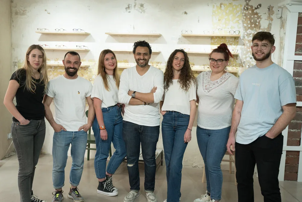 Groupe de sept jeunes adultes posant ensemble dans une pièce avec des étagères à lunettes en arrière-plan.