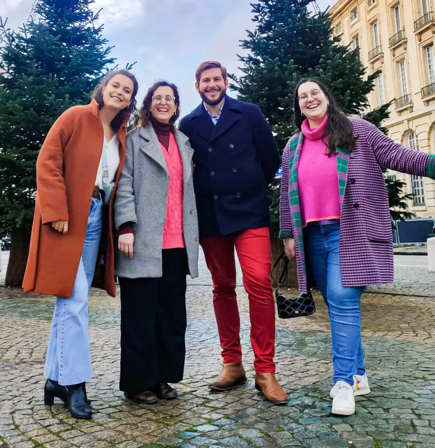Groupe de quatre personnes souriantes posant devant deux sapins de Noël en ville.