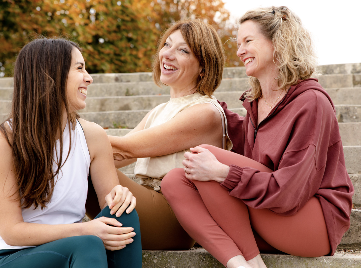 Trois femmes assises sur des marches extérieures, souriant et riant, partageant un moment convivial.