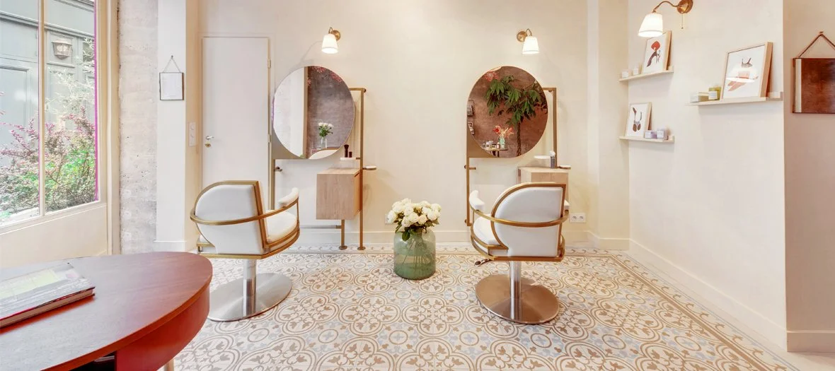Salon de coiffure avec deux chaises, deux miroirs ronds, un vase de fleurs au centre et des décorations murales.